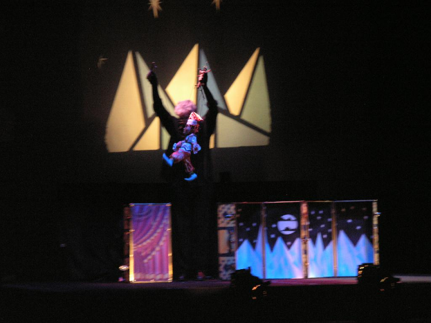 Eine Person auf einer Bühne, die mit Puppen spielt und sie an Fäden hält, mit Dekoration und Lichtern im Hintergrund.