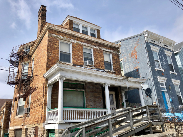 Eine Reihe von baufälligen Häusern an einer Straßenecke mit zerbrochenen Fenstern, einstürzenden Wänden, Geländern, Stufen, einem Zaun, einer Satellitenschüssel, Drähten und einem bewölkten Himmel im Hintergrund.