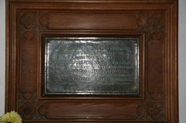 Eine Gedenktafel mit Text ist an einer braunen Wand oder Holztür angebracht, mit einem gelben Objekt, das einer Vase ähnlich ist, in der unteren linken Ecke.