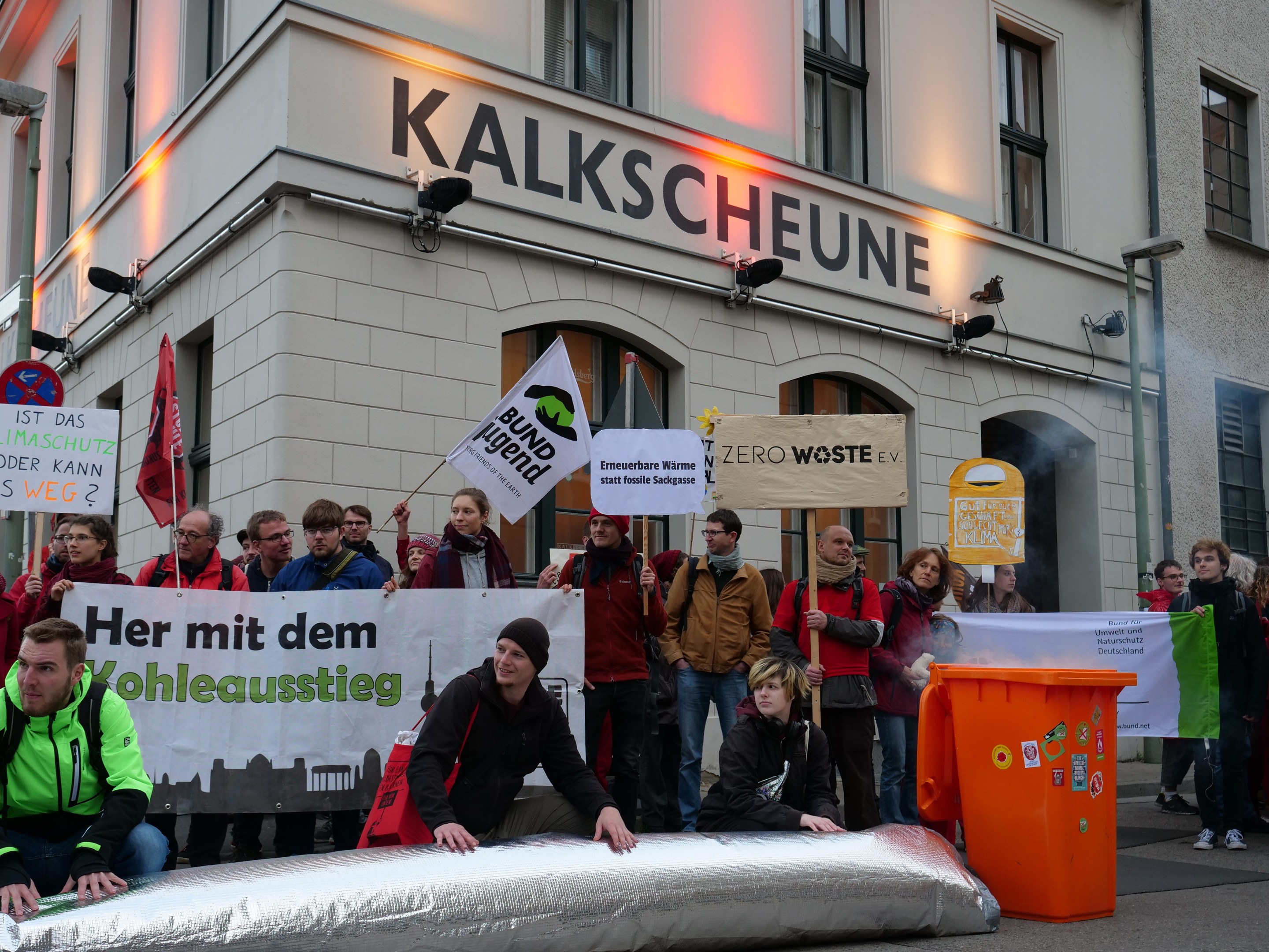 Eine Gruppe von Menschen mit Protestschildern und Transparenten vor einem Gebäude, mit zwei Personen im Vordergrund und einem Müllcontainer rechts.