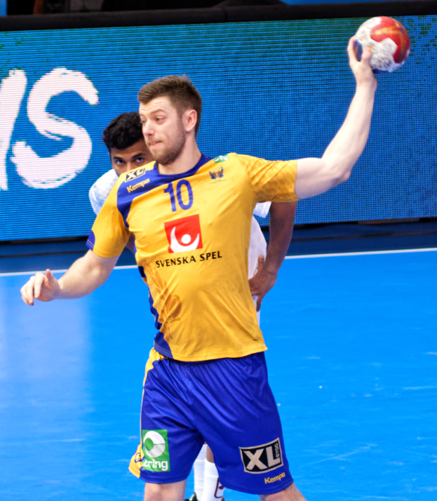 Ein Mann in einem gelben T-Shirt und blauen Shorts, der Handball auf einem Platz spielt und einen Ball hält, mit einem Hintergrundbild, das "Svenský Spel Handball World Cup 2019" anzeigt.
