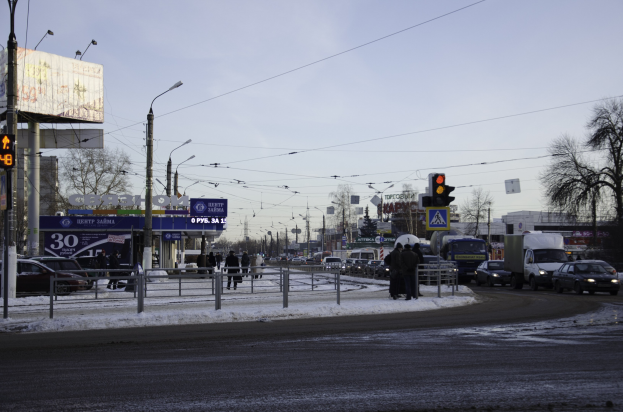 Eine verschneite Stadtstraße mit starkem Verkehr, Straßenlaternen, Fahrzeugen, Geländern, Polen, Schildern, Drähten, Bäumen, Gebäuden und Fußgängern unter einem bewölkten Himmel.