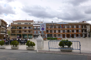 Ein belebter Stadtplatz mit sitzenden und stehenden Menschen, Topfpflanzen, Metallabsperrungen, einem Schild an einem Pfahl, Straßenlaternen mit Flaggen, umliegenden Gebäuden mit Fenstern und einem bewölkten Himmel.