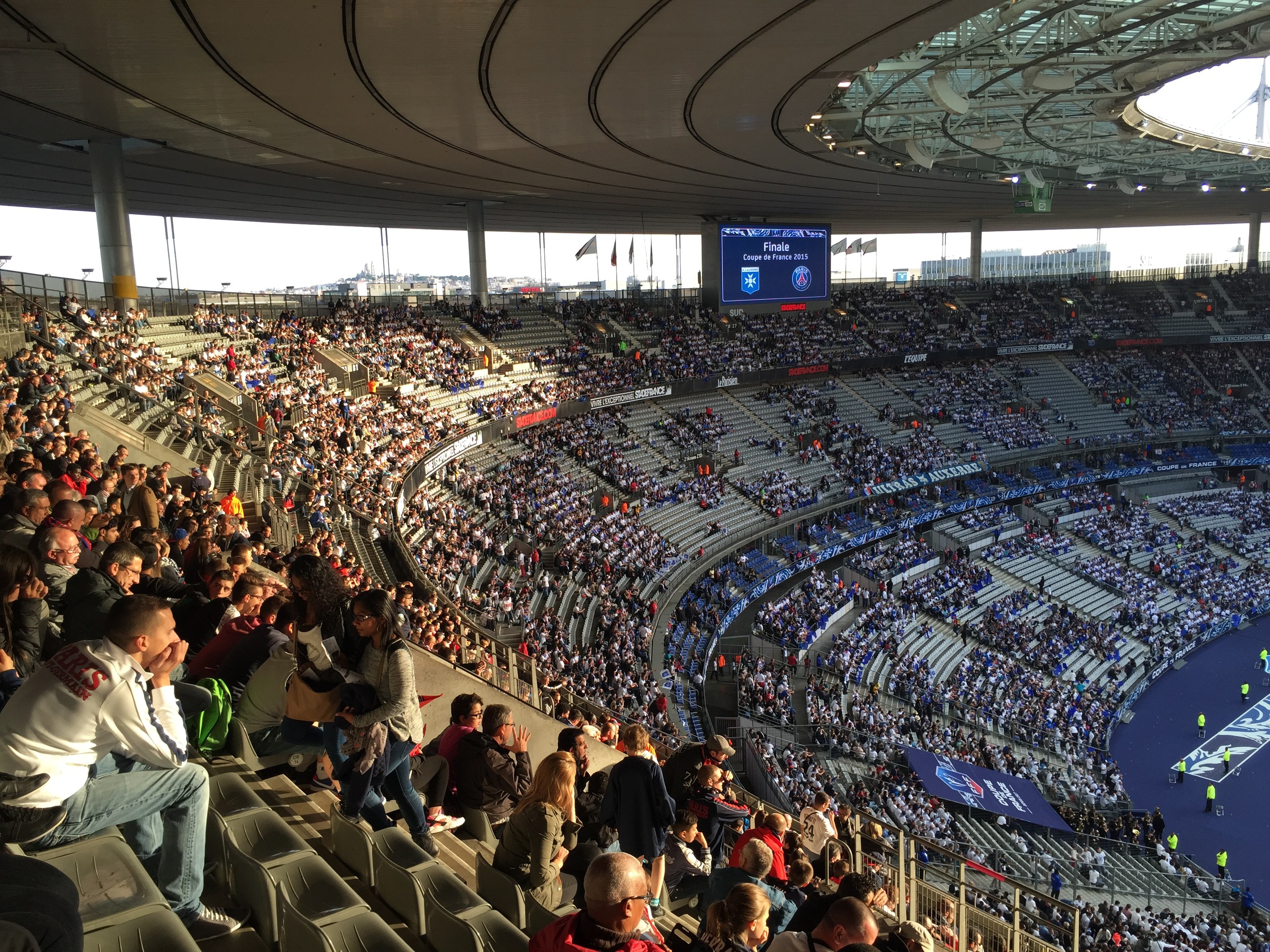 Große Menschenmenge in einem Stadion bei einem Fußballspiel, mit einer Bühne, Fahnen, Pfählen, einem Bildschirm und der Allianz Arena im Hintergrund sichtbar.