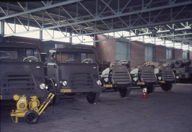 Eine Gruppe von Lkw steht in einem Gebäude mit einem Feuerlöscher auf dem Boden, Metallstangen, Fenstern und einem Dach mit Deckenlampen.