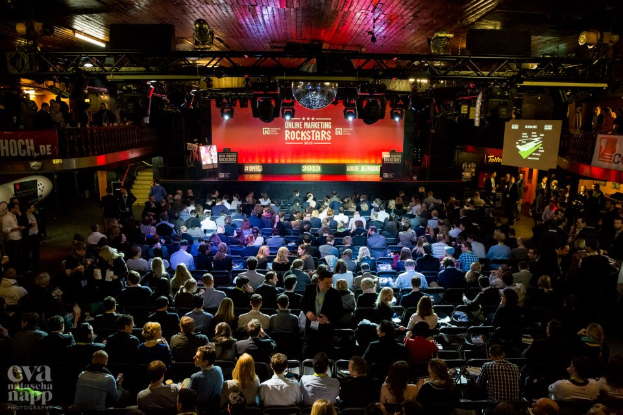 Eine große Menschenmenge sitzt auf Stühlen vor einer Bühne mit Bildschirmen und Bannern, die von Scheinwerfern beleuchtet wird und von Geländern umgeben ist, mit einem Wasserzeichen in der linken unteren Ecke, das eine Atmosphäre der Aufregung und Vorfreude für ein Online-Marketing-Event der Rockstars schafft.