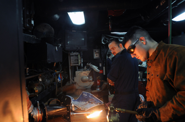 Zwei Männer mit Brille arbeiten an einem Metallstück in einer Werkstatt, einer verwendet ein Schweißgerät, während Werkzeuge und Equipment auf dem Tisch liegen, mit einem Fernseher und Lampen im Hintergrund.