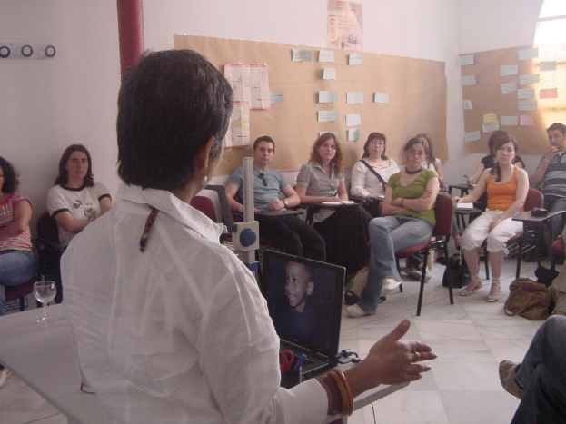 Eine Person in einem weißen Hemd steht an einem Rednerpult und hält eine Gruppe von Menschen in einem Raum mit Plakaten und Papieren auf der Wand an.