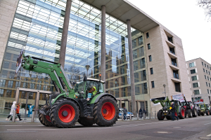 Traktorgruppe fährt eine Straße vor einem Gebäude entlang, mit Menschen auf dem Gehweg und einem Baum auf der rechten Seite während einer Demonstration.