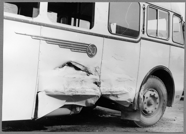 Ein Schwarz-Weiß-Foto eines beschädigten Busses am Straßenrand, dessen Front eingedrückt ist, Scheiben zerbrochen und die Fahrertür offen steht.
