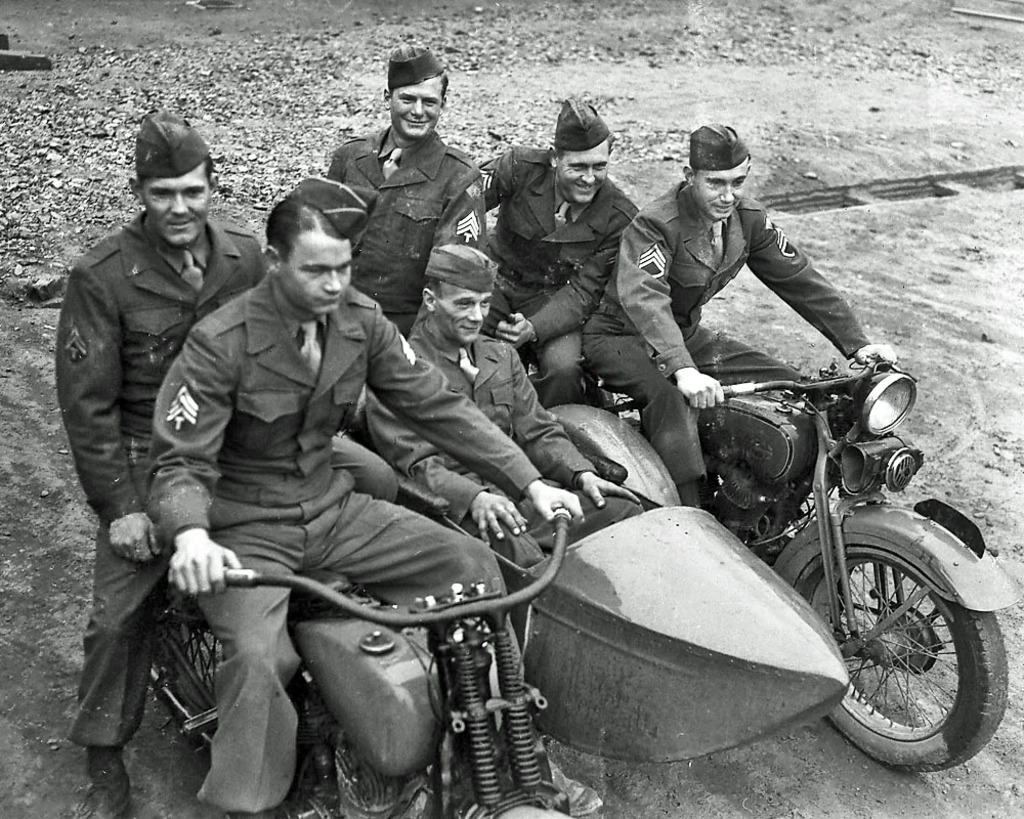Ein Schwarz-Weiß-Foto von mehreren Männern, die auf Fahrrädern sitzen und lächeln, auf einer Straße.