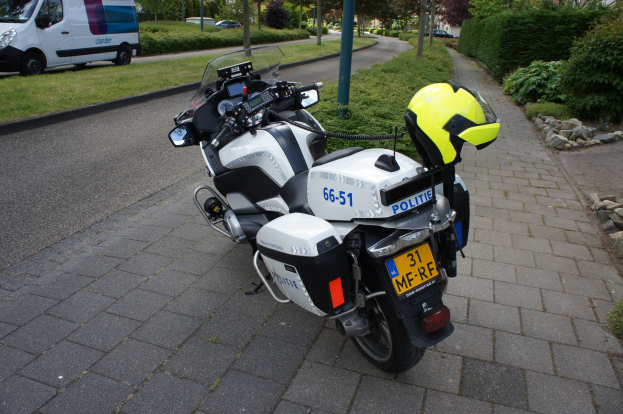 Ein auf der Straße geparktes Polizeimotorrad mit einem Helm darauf, umgeben von Bäumen, Pfählen, Gebäuden, Pflanzen, Gras, Steinen und anderen Fahrzeugen im Hintergrund.