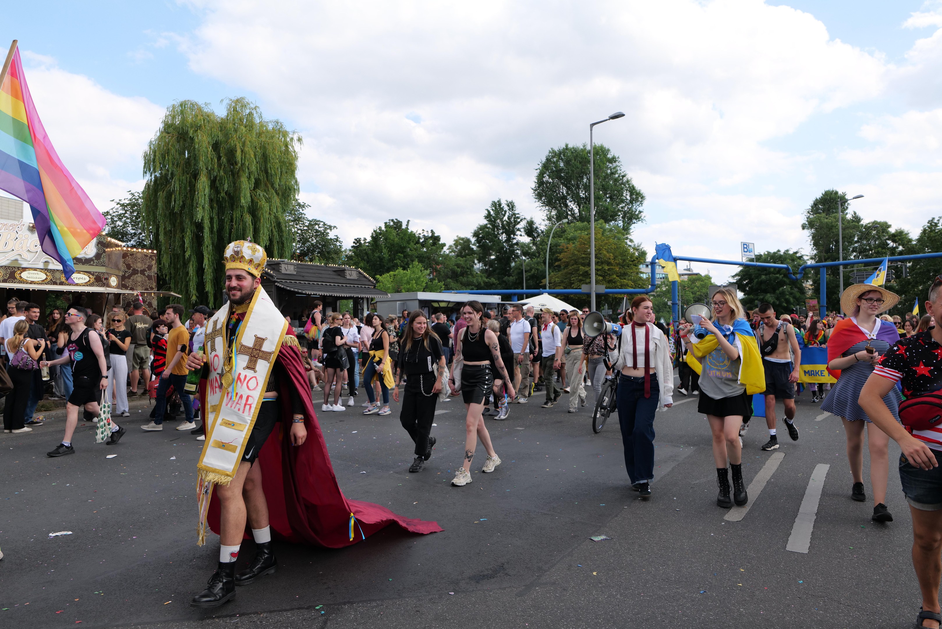 Eine Gruppe von Menschen marschiert bei der Pride Parade 2018 mit einer Regenbogenfahne und Musikinstrumenten, einige tragen Mützen, vor einem Hintergrund aus Laternenmasten, Bäumen, Schuppen und einem bewölkten Himmel.