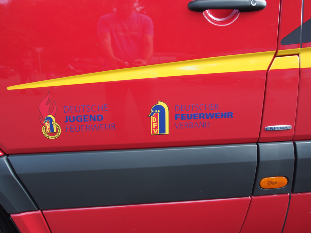 Ein roter Van mit einer gelben Streifen, einem Griff und einem Logo mit einem Löwen in einem Schild, umgeben von einem Lorbeerkranz, mit der Aufschrift "Deutscher Jugendfeuerwehrverband".