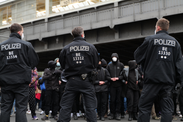 Eine Gruppe von Polizisten in Uniform steht vor einer Menge von Menschen in schwarzen Uniformen und Masken mit einer Brücke und einem Gebäude im Hintergrund während einer Demonstration in einer Stadt.