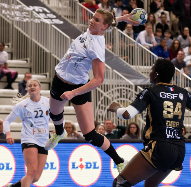 Eine Gruppe von Frauen, die auf einem Court Handball spielen, mit einem Ball in der Luft, Zuschauern im Hintergrund und einer "Handball-Weltmeisterschaft 2019 - Tag 2"-Tafel.
