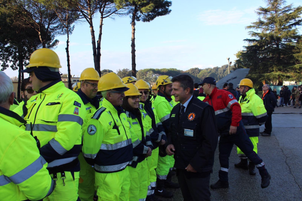 Eine Gruppe von Menschen in gelben Westen und Helmen, einige mit Mützen, steht zusammen auf einer Straße, umgeben von Bäumen unter einem klaren blauen Himmel.