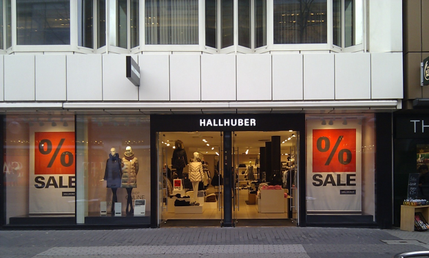 Ein Hallhuber-Laden mit Schaufensterpuppen, Kleiderbügeln, Gegenständen auf Tischen, Schildern, einem Metallzaun und einer Decke mit Deckenleuchten.