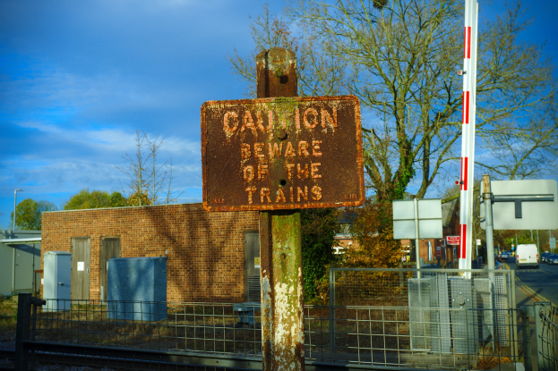 Warnschild an einem Bahnübergangszaun mit Bäumen, Strommasten, einem Gebäude, Containern, Fahrzeugen auf einer Straße und einem bewölktem Himmel.