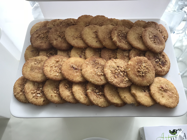 Ein weißer Teller voller Kekse steht auf einem Tisch umgeben von Gläsern und anderen Gegenständen, mit einem Blatt Papier am Boden mit Text und Logo.