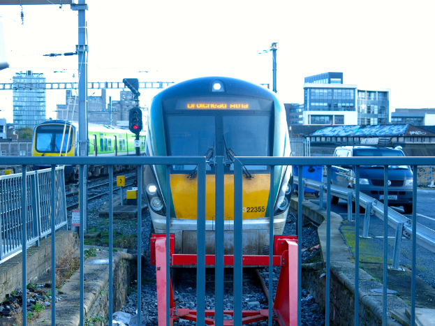Zwei Züge auf dem Gleis mit einem Zaun im Vordergrund, ein Auto auf der Straße rechts, eine Signalleuchte auf einem Pfahl, Gebäude im Hintergrund und der Himmel darüber.