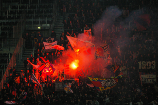 Eine große Menschenmenge in einem Stadion mit Fahnen, Bannern und einem zentralen Feuer, umgeben von Stufen und Geländern.