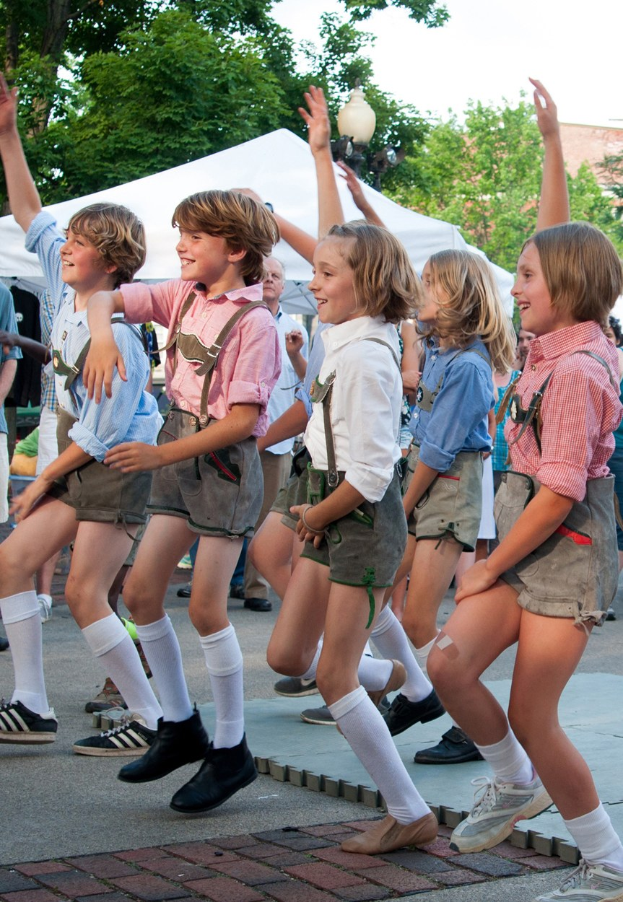 Kinder in traditioneller bayrischer Kleidung tanzen auf einer Straße während eines Outdoor-Events, umgeben von einem Publikum, Zelten und Gebäuden unter einem klaren blauen Himmel.