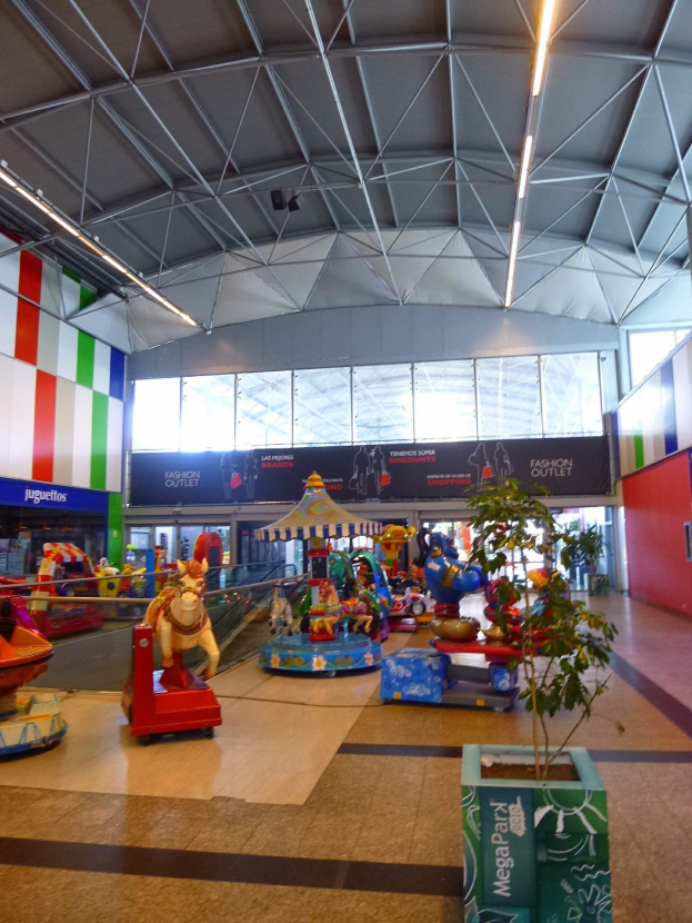 Großes Indoor-Vergnügungspark-Areal in einem Einkaufszentrum mit verschiedenen Fahrgeschäften, einem Topf mit einer Pflanze, verstreuten Spielzeugen, Informationsschildern, Deckenleuchten und Metallstützen.