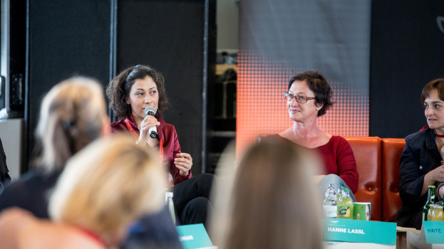 Eine Gruppe von Frauen sitzt vorn, wobei eine links ein rotes Kleid trägt und ein Mikrofon hält, und im Hintergrund eine Roll-up-Banner und eine schwarze Plane.