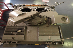 Ein großer Militärpanzer mit sichtbaren Ketten und Turm ist auf einem Boden positioniert, mit einem Metallgeländer auf der linken Seite.