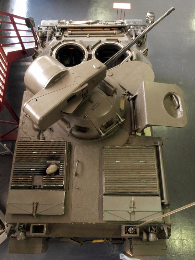 Ein großer Militärpanzer mit sichtbaren Ketten und Turm ist auf einem Boden positioniert, mit einem Metallgeländer auf der linken Seite.