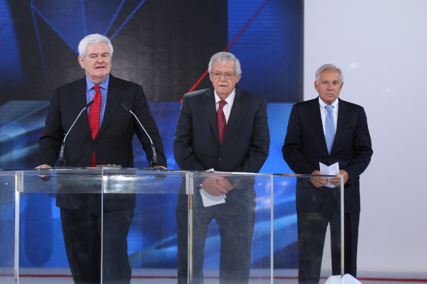 Ein älterer Mann in einem schwarzen Anzug und roter Krawatte steht an einem durchsichtigen Rednerpult auf einer Bühne, mit einem blauen und weißen Banner im Hintergrund.