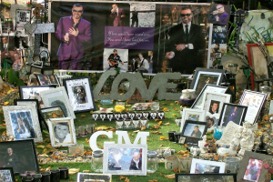 Ein provisorisches Mahnmal mit Fotos, Kerzen, Blumen und anderen Gegenständen auf Gras vor einem Gebäude mit Bäumen und einer Fahne im Hintergrund.