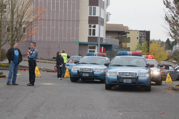 Autos auf einer Straße mit vier Menschen in der Nähe, Gebäude mit Fenstern im Hintergrund, Bäume und Notfallwarnkegel.