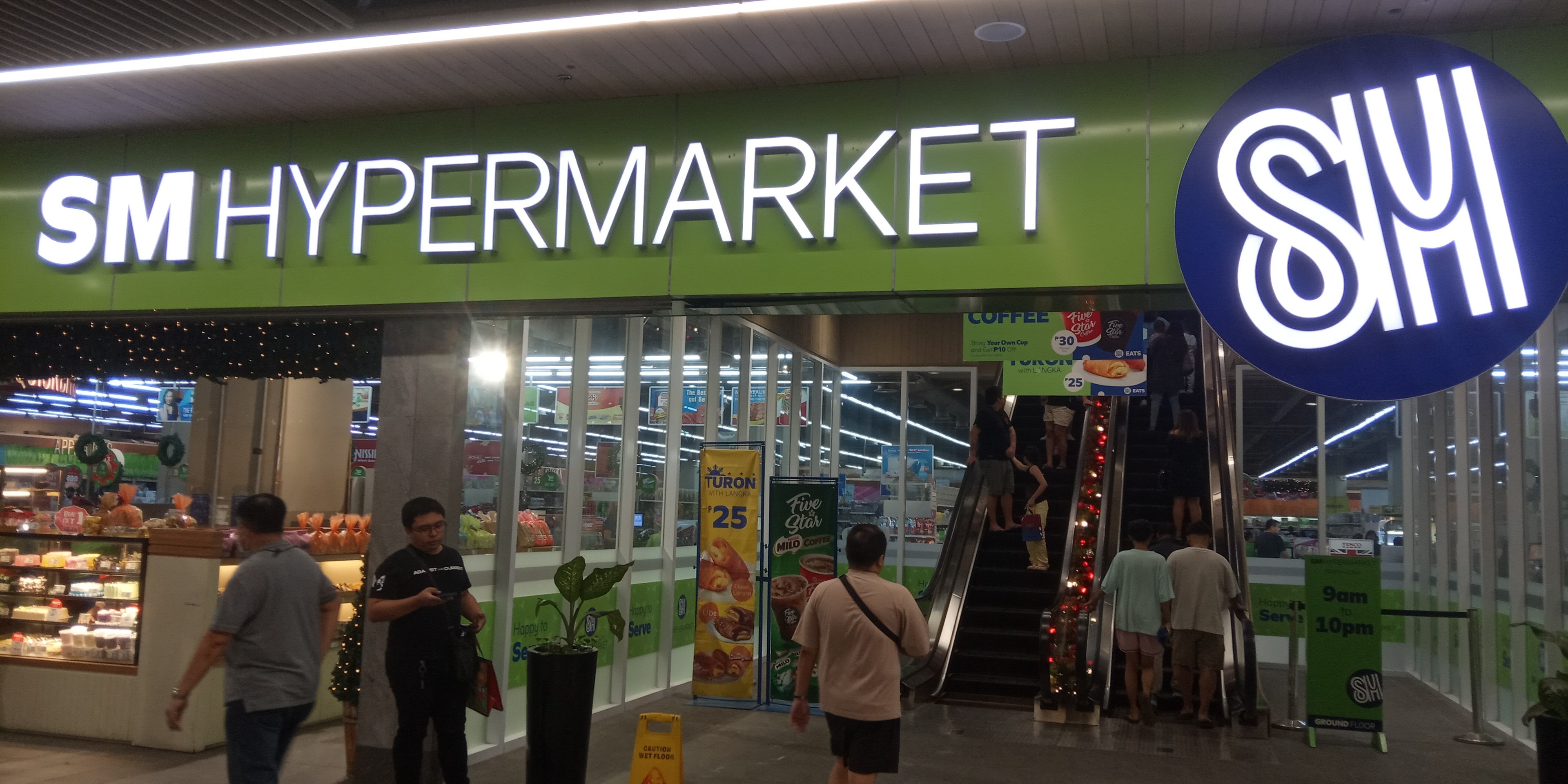 Eine Gruppe von Menschen vor einem hell erleuchteten "SM Hypermarket"-Geschäft mit Deckenleuchten, Sichttafeln und Zimmerpflanzen.