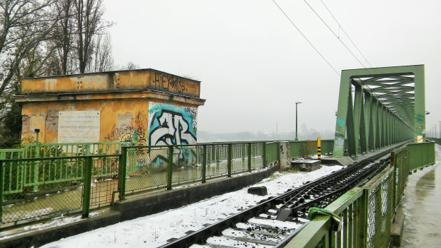 Eine schneebedeckte Zuggleis mit Graffiti an der benachbarten Wand, umgeben von Gelöndern, Pfosten, Lichtern, Kabeln und Überirdischen Leitungen, mit Bäumen und einem sichtbaren Himmel im Hintergrund.