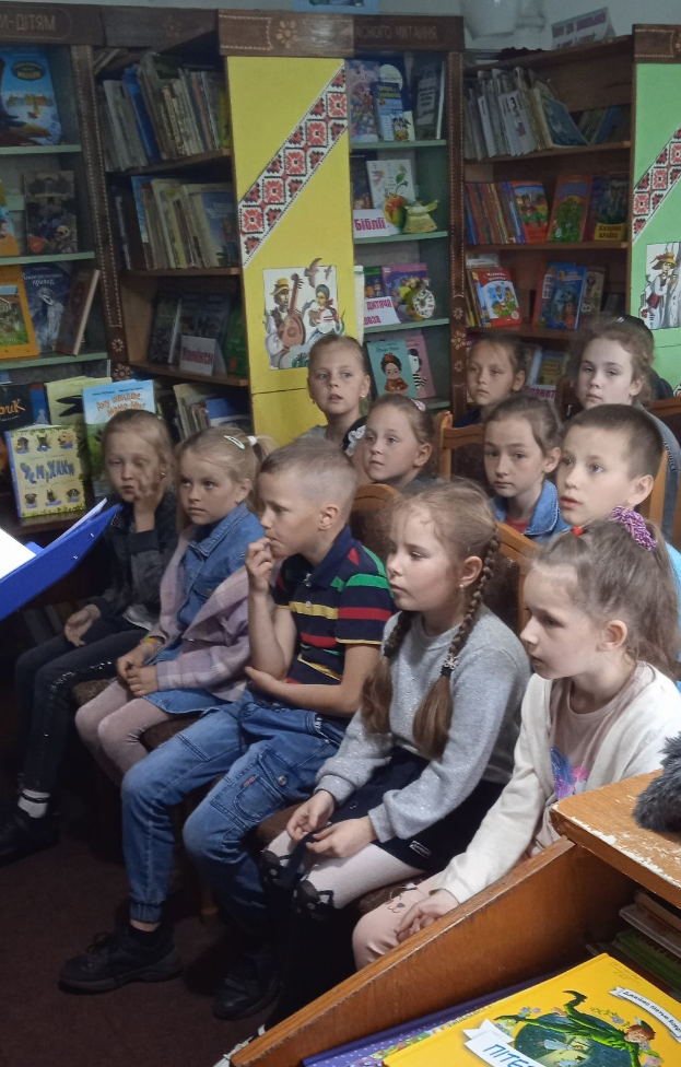 Eine Gruppe von Kindern sitzt in einer Bibliothek umgeben von Bücherregalen, mit einigen Lesen an Tischen auf beiden Seiten.