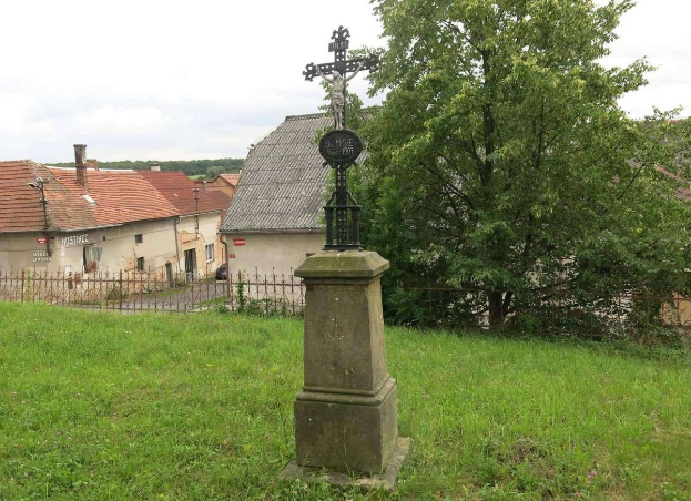 Ein Kreuzdenkmal auf einer Wiese, umgeben von einem Metallzaun, Bäumen, Häusern und einem bewölktem Himmel, gewidmet den Holocaust-Opfern.