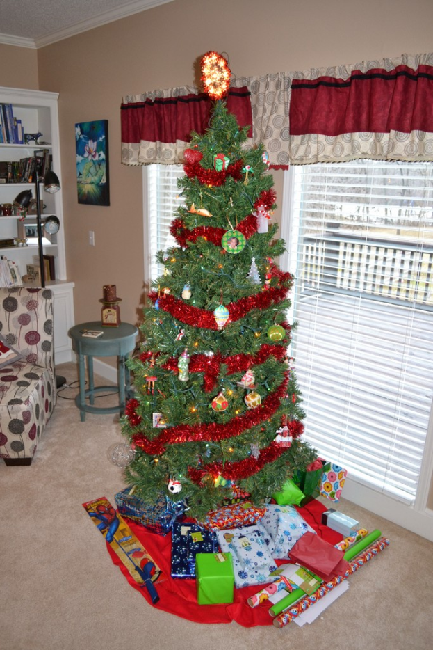Ein Weihnachtsbaum mit Geschenken davor, eine Couch, ein Tisch, ein Bücherregal, eine Lampe mit Stange, ein Bild an der Wand und Fenster mit Vorhängen.