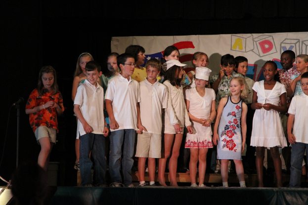 Mehrere Kinder stehen auf einer Bühne, zwei in der Mitte mit Mützen, ein Mikrofonständer links und ein Banner im Hintergrund.