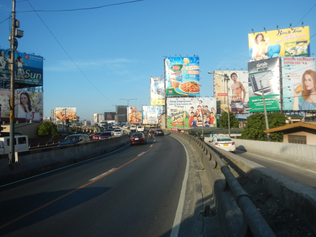 Eine vielbefahrene Autobahn mit mehreren Fahrspuren, hohe Werbetafeln, Strommasten mit Leitungen, Bäume am Straßenrand und ein klarer blauer Himmel.