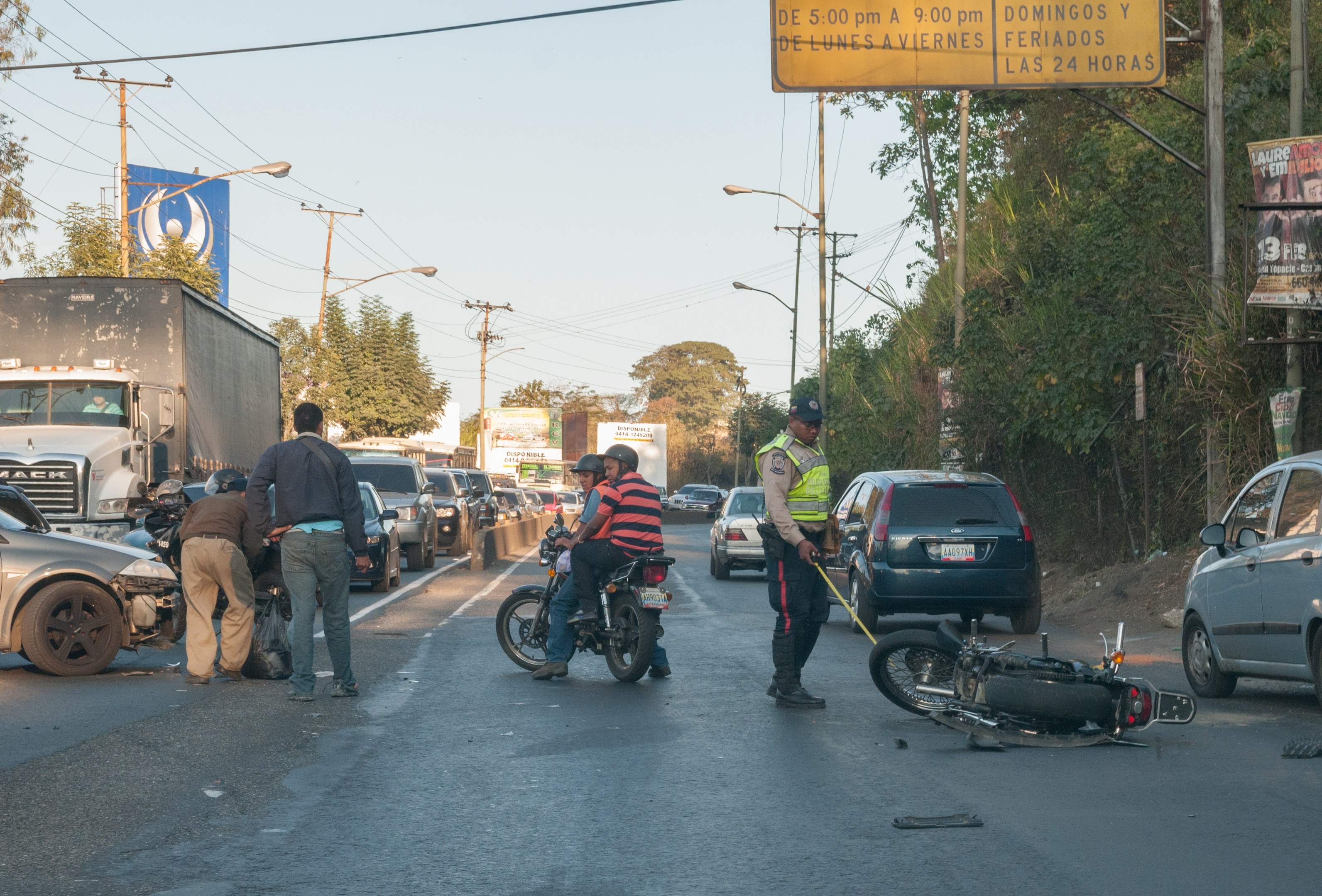 Eine Gruppe von Menschen steht um ein verunglücktes Motorrad auf der Straße herum, mit mehreren Fahrzeugen, darunter ein Lastwagen, und Hintergrundelementen wie Bäumen, Pfählen, Lampen, Schildern und dem Himmel.