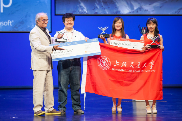 Vier Personen auf einer Bühne, jede hält Auszeichnungen, Schecks und eine Fahne, lächeln, mit einem digitalen Bildschirm im Hintergrund.