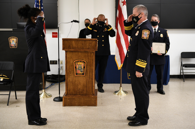 Eine Gruppe uniformierter und maskierter Personen steht vor einem Podium mit einem Mikrofon, umgeben von Fahnen, Stühlen und Tischen, während einer Vereidungszeremonie für einen neuen Feuerchef, wobei eine Person ein Zertifikat hält.