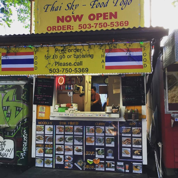 Ein Schaufenster mit verschiedenen Artikeln wie Getränkedosen, Speisekarten, einer Uhr, Gewürzen in Plastikbehältern, Kondimente, Werbetafeln und Namensschildern vor einem Himmel.