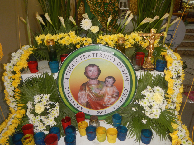 Ein mit bunten Blumen, Kerzen und einer Tafel mit einem Porträt und Text geschmückter Tisch, vor einer Kulisse aus Pflanzen, Stühlen und anderen Gegenständen, die eine festliche Atmosphäre für die Bruderschaft des Heiligen Josef schafft.