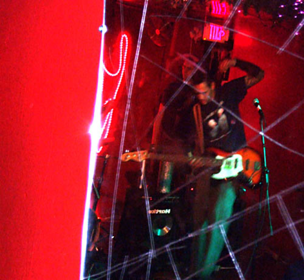 Ein Mann in einem schwarzen T-Shirt steht in der Mitte und hält eine Gitarre, während ein anderer Mann hinter ihm möglicherweise Instrumente spielt, unter hellen Scheinwerfern und vor einer roten Wand auf einem Musikkonzert.