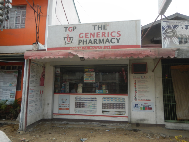 Außenansicht der "Generics Apotheke" mit umliegenden städtischen Elementen wie Gebäuden, Fahrzeugen und Infrastruktur unter einem klaren blauen Himmel.