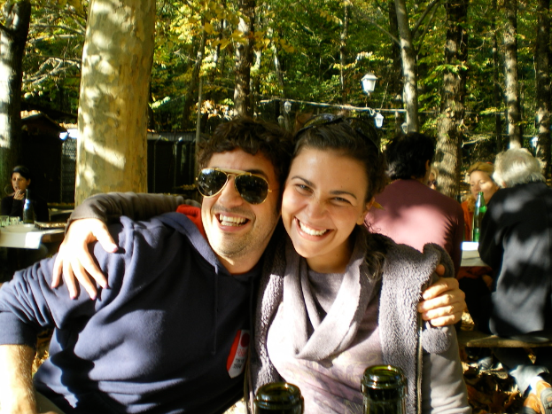 Ein Paar sitzt im Vordergrund, umarmt und lacht, während Menschen an Tischen mit Flaschen im Hintergrund sitzen, mit Bäumen und Laternen im Hintergrund.