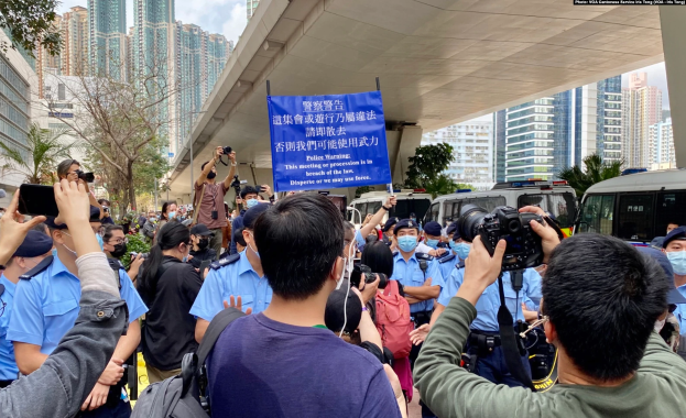 Eine Gruppe von Menschen steht vor einer Menge, einige halten Kameras und Telefone, mit einem Banner "Hong Kong Police Wants to Stop the Violence Against Women", Fahrzeuge, Bäume, Gebäude und ein bewölkter Himmel im Hintergrund.